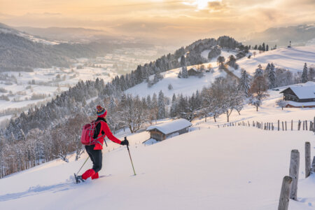 Une randonneuse traverse un champ enneigé en raquettes, avec un paysage alpin enneigé et des chalets de bois en arrière plan