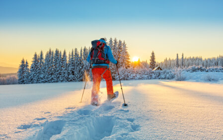 Un homme marche en raquettes au milieu d'un paysage de montagne totalement enneigé, avec un coucher de soleil en fond